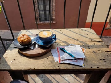 Blue coffee cup and pastry on a sunlit wooden table with postcards and a pen